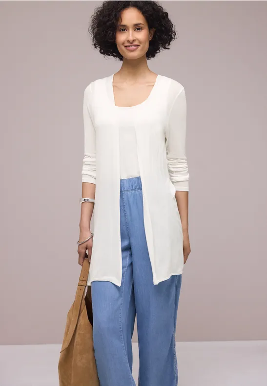 Eine Person trägt ein weißes, offenes Cardigan mit drei-viertel Ärmeln, blaue weite Hose und eine beige Tasche.