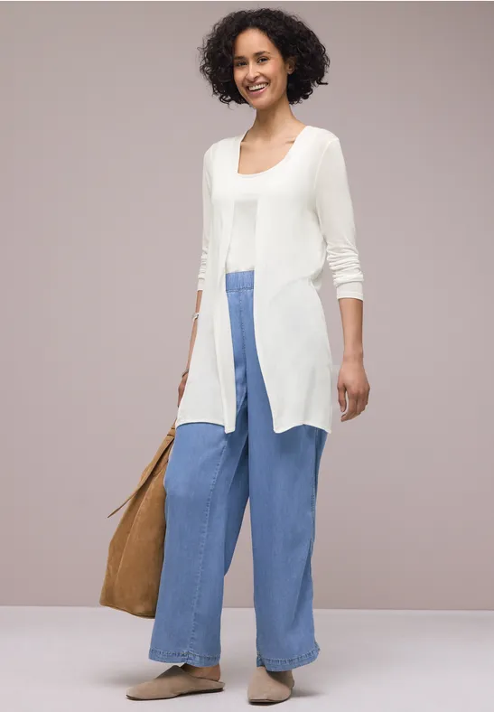Eine Person trägt eine weiße, lange Strickjacke, weite blaue Hosen, beiges Espadrille-Paar und eine braune Tasche.
