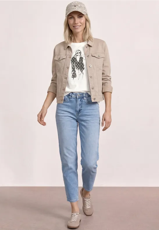 Frau in weißem T-shirt mit Muster, beige Jacke, hellblauen Jeans und Sneakern vor hellem Hintergrund.