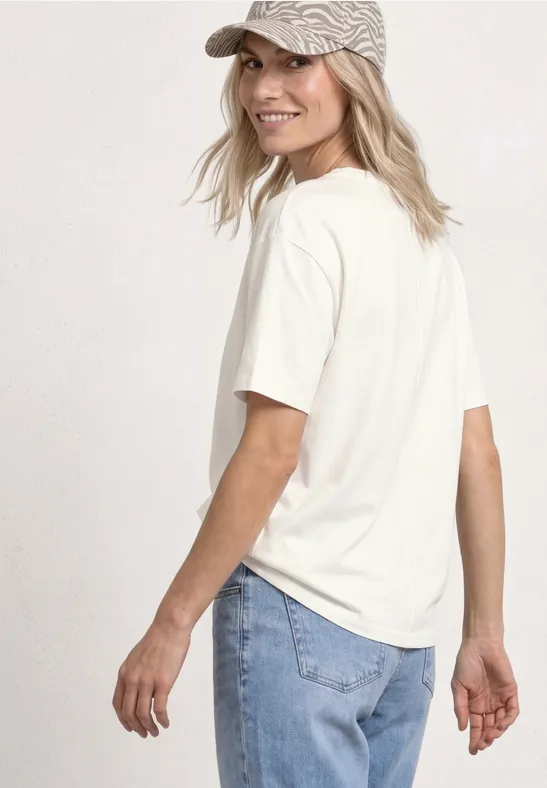 Person mit Baseballcap, weißem T-Shirt und Jeans steht seitlich und blickt weg.