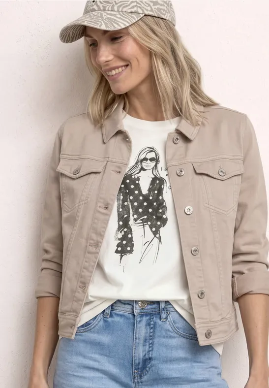 Frau mit beiger Jacke, weißem T-Shirt mit Fashion-Illustration, blauen Jeans und Baseballcap vor hellem Hintergrund.