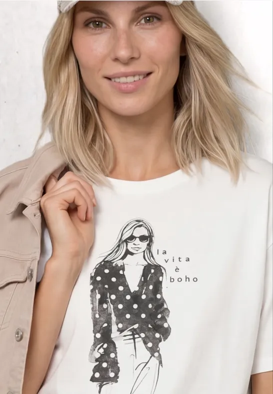 Blonde Frau in weißem Boho-Shirt mit modischem Print, die eine Jacke über der Schulter hält.