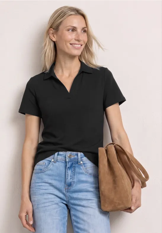 Femme portant un polo noir, un jean bleu et un sac marron devant un fond clair.