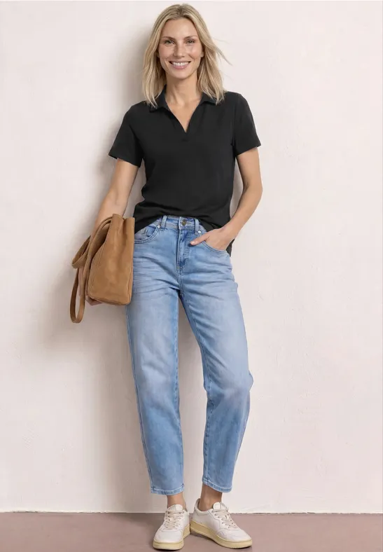Femme en polo noir, jean et baskets blanches, debout devant un mur clair avec un sac beige sur l'épaule.