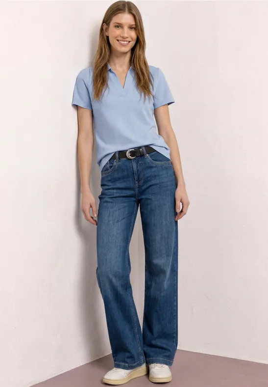 Une femme porte un polo bleu clair, un jean bleu et des baskets blanches, posant devant un mur neutre.
