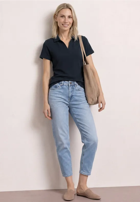 Femme debout devant un fond clair, portant un polo noir, un jean bleu, des mocassins bruns et un sac en bandoulière.
