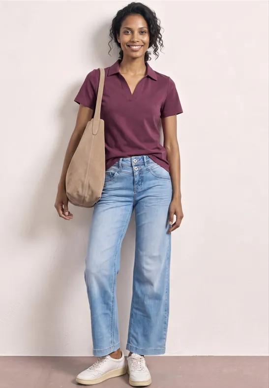 Person trägt lila Poloshirt, helle Jeans, weiße Sneakers und trägt eine beige Stofftasche.