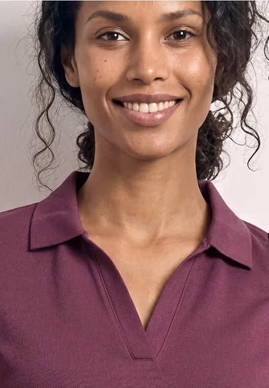 Eine Person mit lockigen Haaren trägt ein purpurfarbenes Poloshirt. Nur Oberkörper und Hals sind zu sehen.