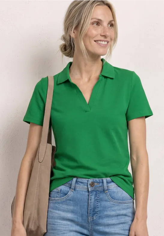 Femme en polo vert et jean avec un sac beige sur l'épaule, buste photographié de face.