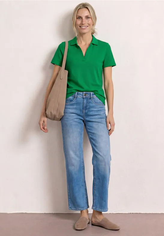 Femme en polo vert, jeans, sac beige et mocassins devant mur clair.