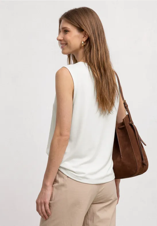 Vrouw in wit mouwloos topje en beige broek, draagt een bruine schoudertas, kijkt opzij.