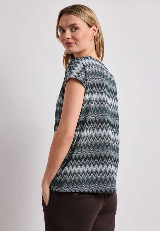 Vrouw van opzij in een grijs-groen-zwart zigzagtop met korte mouwen en een donkere broek.