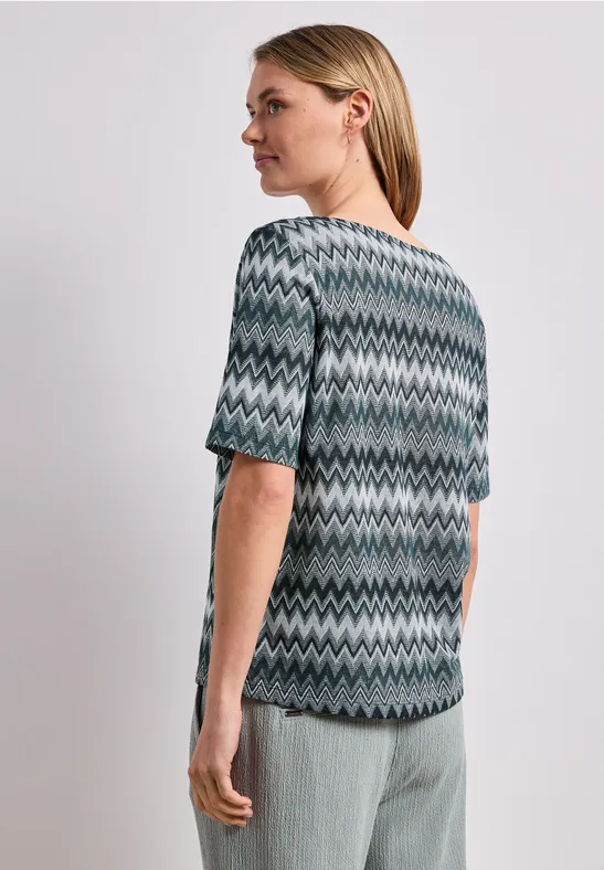 Vrouw van achteren in blauwgrijs zigzagshirt en lichtgrijze broek.