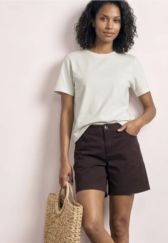 Femme en t-shirt blanc et short marron, tient un sac tressé en paille, fond clair.