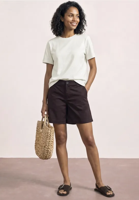 Femme en t-shirt blanc, short noir et sandales noires, tenant un sac en paille.