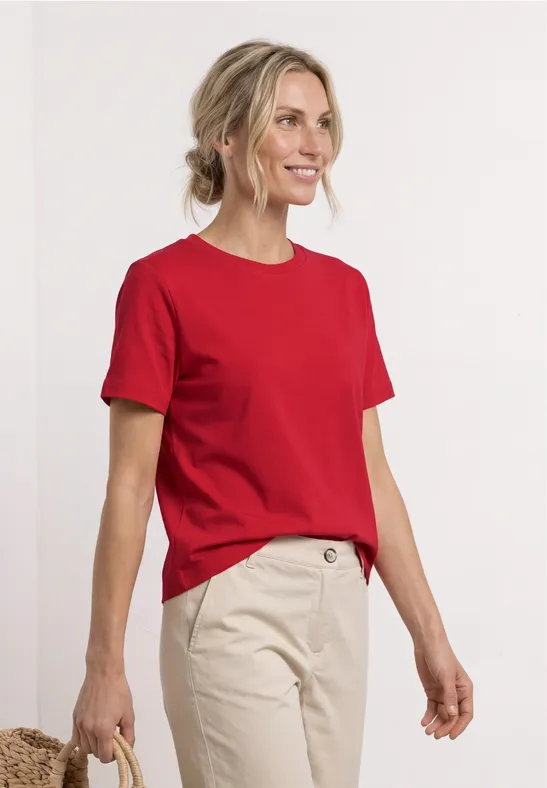 Personne portant un t-shirt rouge et un pantalon beige devant un fond blanc, visage flou.