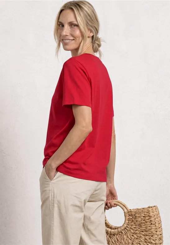 Femme de dos en t-shirt rouge et pantalon beige, tenant un panier tressé, fond clair