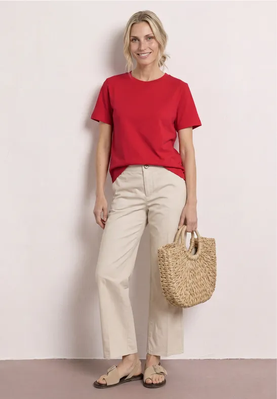 Personne en t-shirt rouge, pantalon beige et sandales, tenant un sac en osier devant un fond clair.