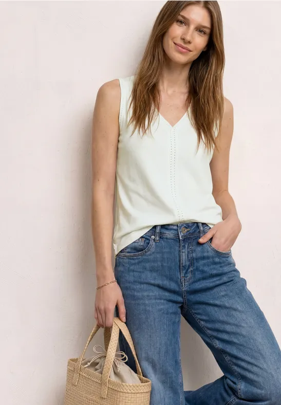 Femme en top blanc à col en V, jean bleu et sac en paille sur fond neutre.