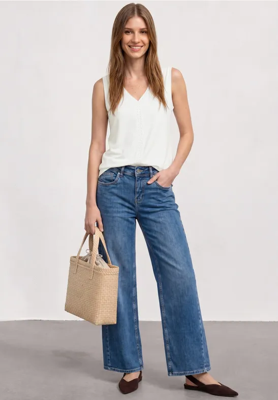 Femme en top blanc, jeans larges et ballerines noires, tenant un sac en paille devant un fond gris.