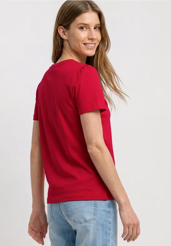 Vrouw in rood T-shirt en blauwe jeans van achteren, hoofd zijwaarts gedraaid.