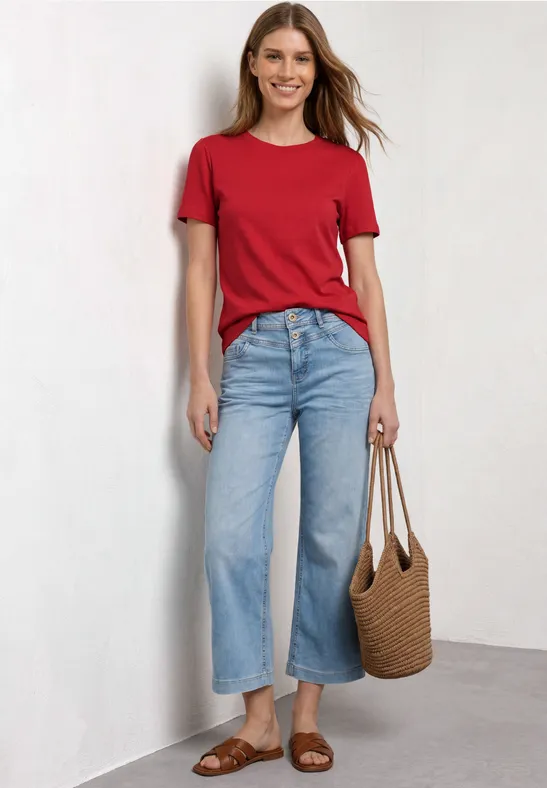 Vrouw in rood T-shirt, wijde blauwe jeans, bruine sandalen en rieten tas voor een witte muur.