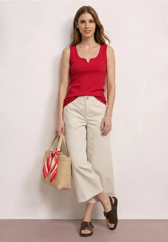 Vrouw in rood topje, beige broek en rieten tas voor een neutrale achtergrond.