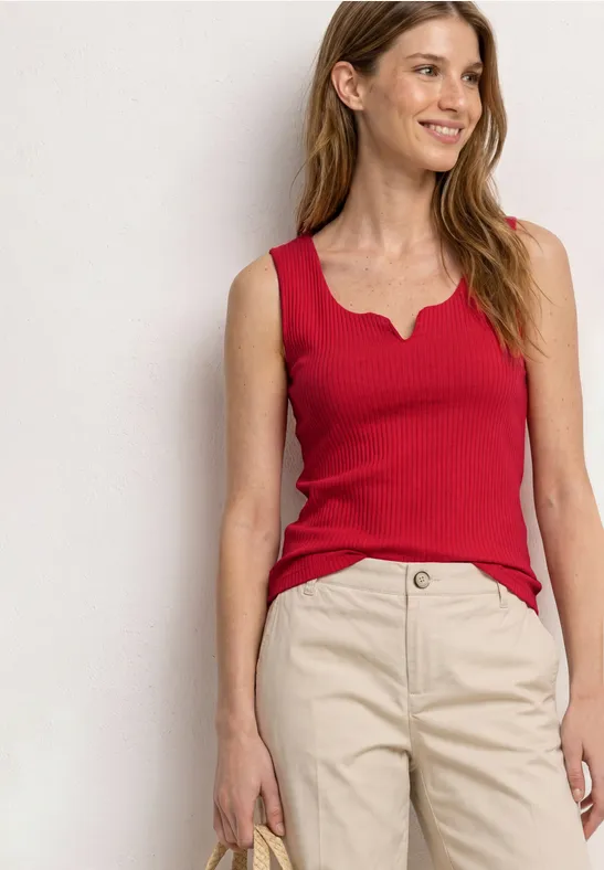 Vrouw staand in een rood mouwloos topje en beige broek.