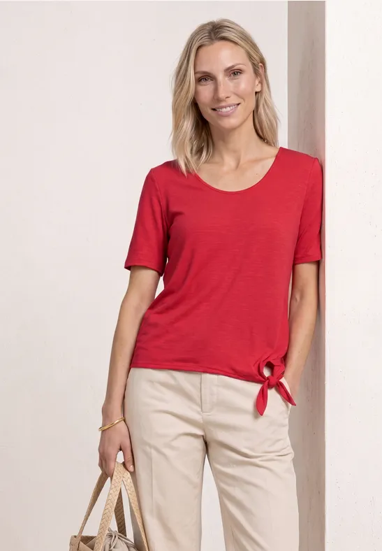 Vrouw in rood t-shirt met knoop, lichte broek en een gevlochten tas.