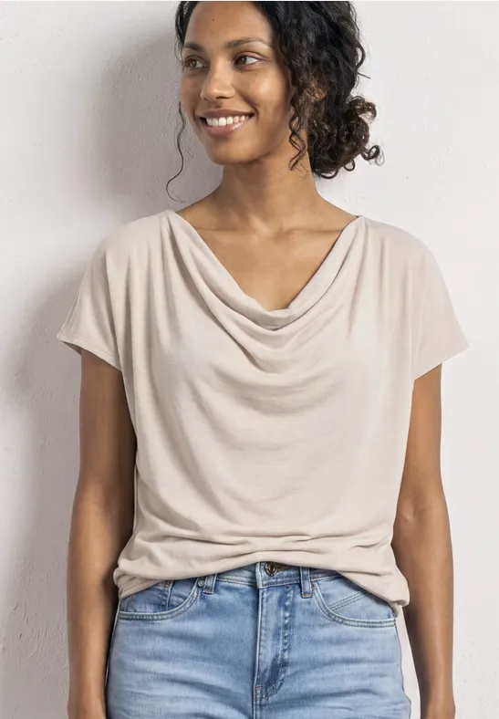 Eine Person trägt ein cremefarbenes Drape-Shirt mit kurzem Ärmel und blaue Jeans vor hellem Hintergrund.