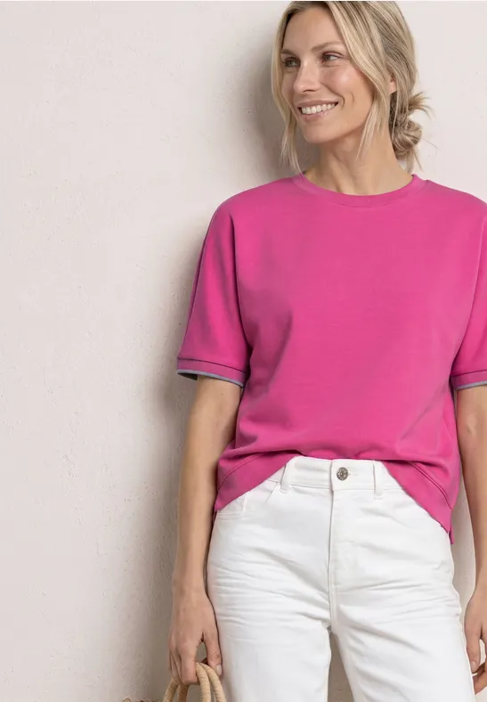 Vrouw in felroze shirt en witte jeans, met klein tasje, neutrale achtergrond.