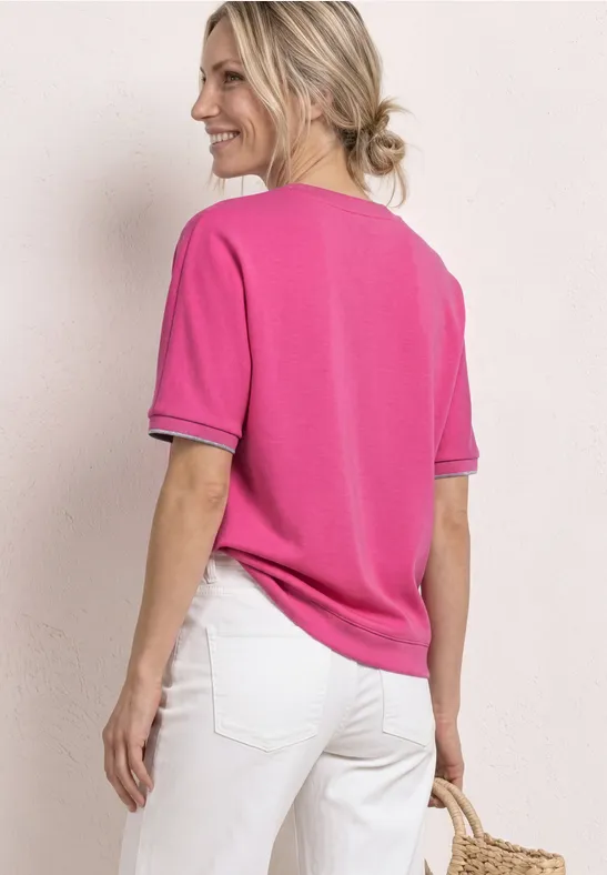 Rugaanzicht van een vrouw in roze shirt met korte mouwen en zwarte rand, witte broek en gevlochten strotas.