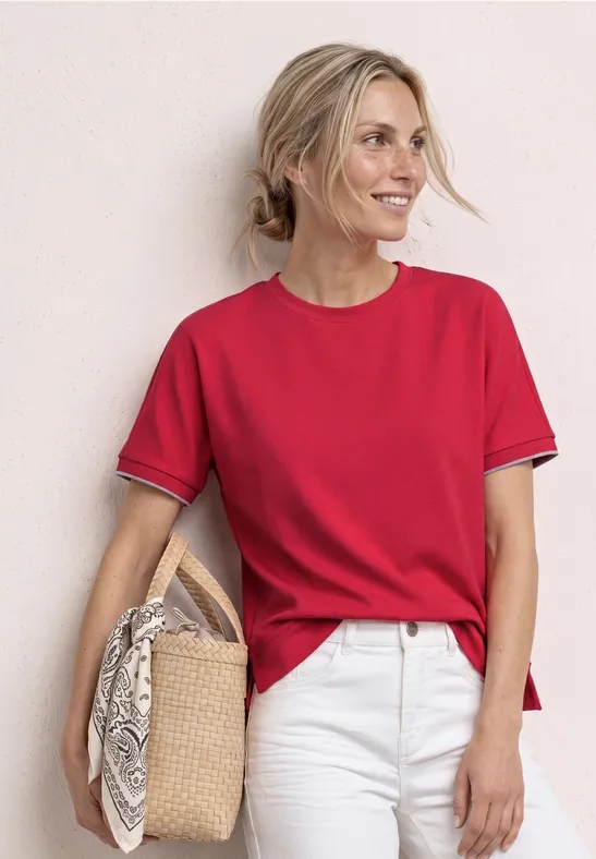 Persoon draagt een rood T-shirt, witte broek en houdt een strooien tas voor een neutrale achtergrond.