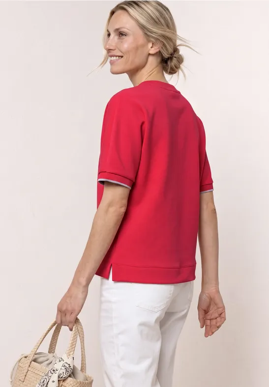Vrouw van achteren in rood shirt met korte mouwen, witte broek en rieten tas in de hand.