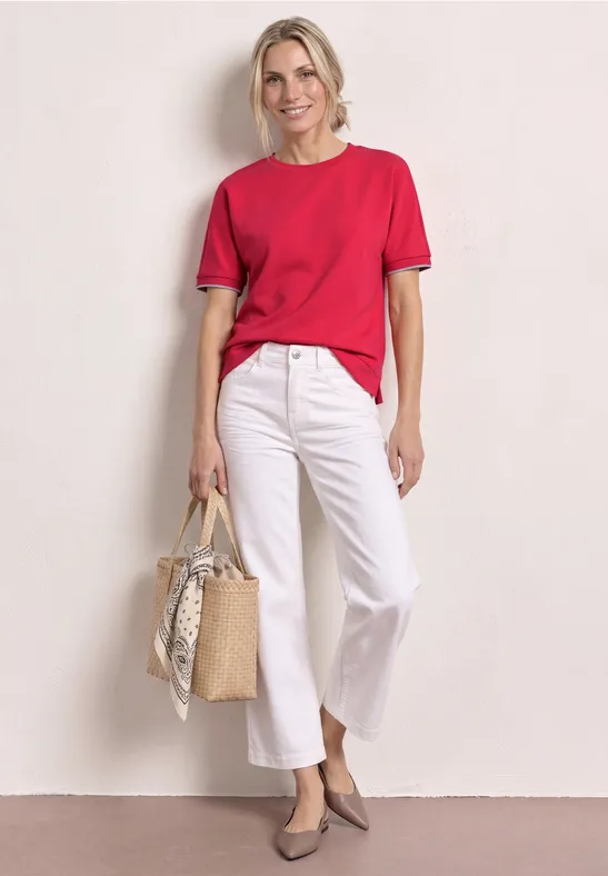 Vrouw met rood T-shirt, witte broek, grijze pumps en een gevlochten strooien tas met sjaal.