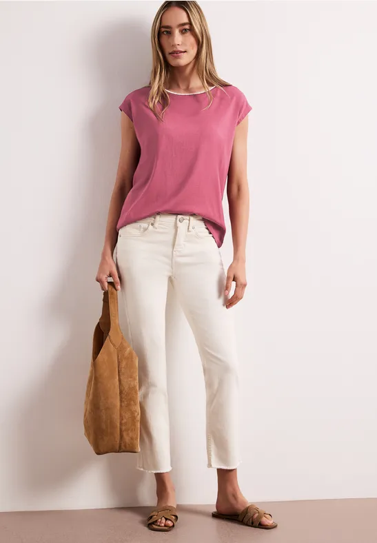 Een vrouw in een roze top, witte cropped jeans, sandalen en bruine tas voor een lichte achtergrond.