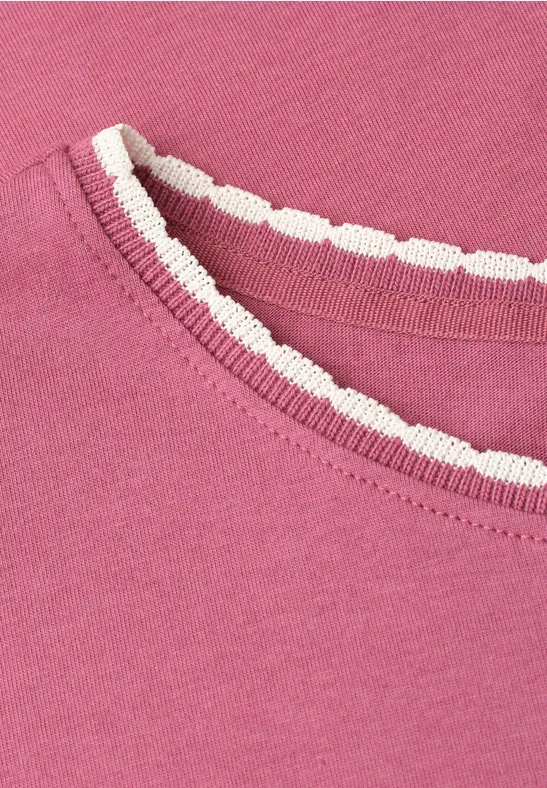 Close-up van een roze T-shirt met een wit, gegolfd biesje bij de halsuitsnijding.