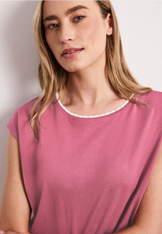 Een persoon draagt een mouwloze roze top met witte kraag en staat voor een witte muur.