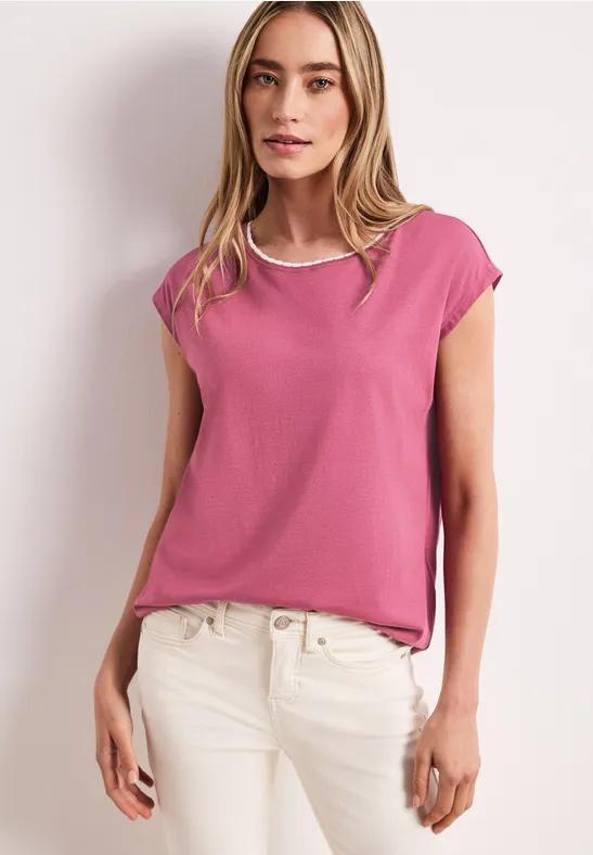 Vrouw in een roze shirt en witte jeans tegen een lichtgrijze achtergrond.