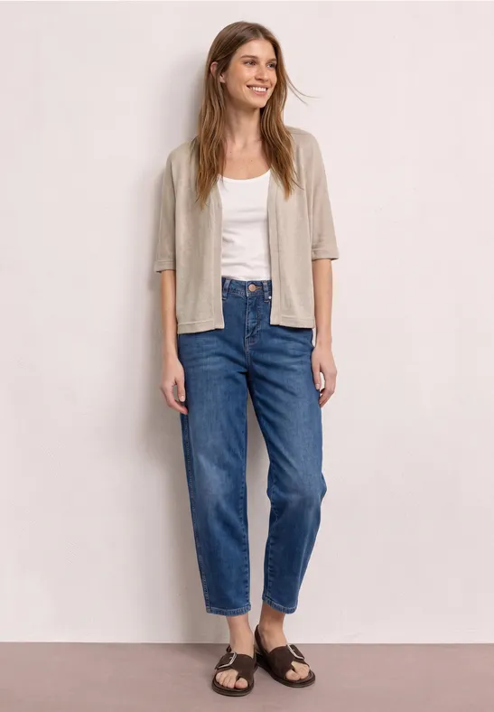 Vrouw met onscherp gezicht voor neutrale achtergrond, in wit top, beige vest, blauwe jeans en bruine sandalen.
