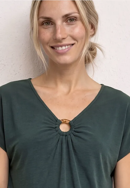 Frau mit grünem T-Shirt und dekorativem Ring am Ausschnitt vor heller Wand.