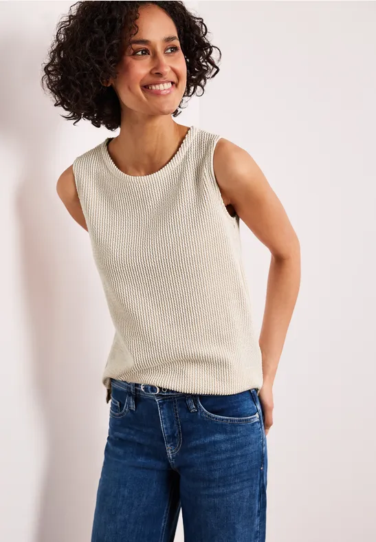 Person steht vor weißem Hintergrund, trägt ärmelloses beiges Oberteil und blaue Jeans.