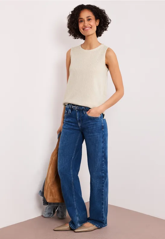 Frau in cremefarbenem Top und weitem Jeans steht lässig vor Wand, Hand in Tasche.