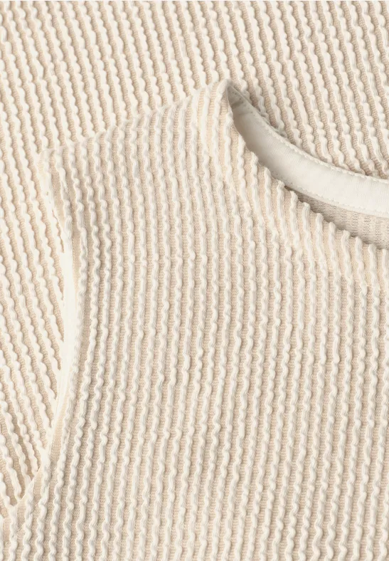 Naheaufnahme eines cremefarbenen, gerippten Strickoberteils mit Rundhalsausschnitt.