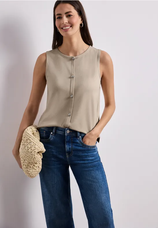 Une femme porte un top sans manches beige et un jean bleu, tenant une pochette tressée.