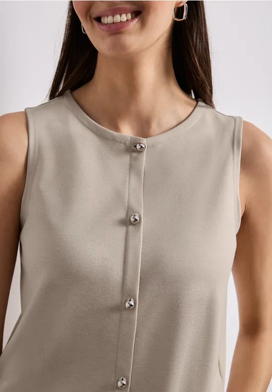 Une femme porte un haut sans manches beige à boutons argentés et sourit.