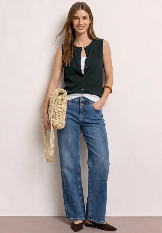 Femme portant un débardeur noir, un t-shirt blanc, jean large, mocassins et sac en osier, devant un mur clair.