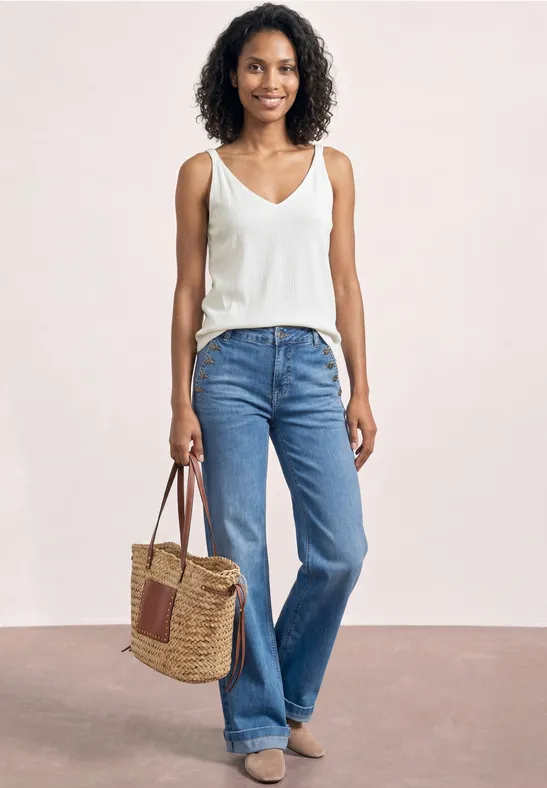 Une femme porte un débardeur blanc, un jean bleu ample et tient un sac en osier tressé.