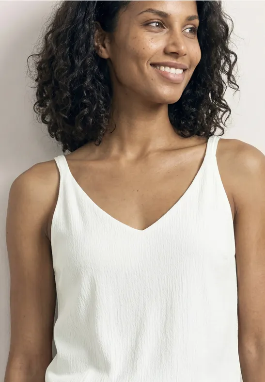 Une personne porte un top blanc sans manches, cheveux bouclés, visage flouté.