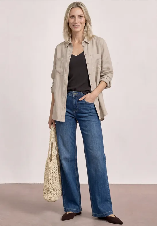 Vrouw met beige overshirt, zwart topje, wijde jeans, gehaakte tas en donkere schoenen.